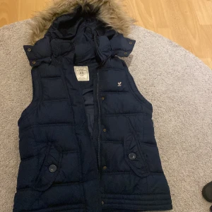 Mörkblå dunväst från H&M L.O.G.G. - Snygg mörkblå dunväst från H&M L.O.G.G. med avtagbar huva och fuskpäls. Västen har quiltad design, två fickor med knappar och dragkedja framtill. 