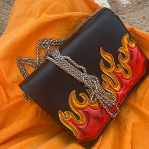 Unik flame-väska🔥🔥 - Cool väska i svart läderimitation med flammor på framsidan och reglerbar axelrem i form av kjedja. 3 stora fack på insidan, 2 med kjedja. Väskans storlek är 18x24cm och är köpt i butik i Belgien på ”sacha iconic”🖤