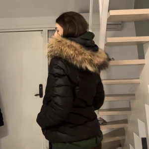 Svart dunjacka med päls - Dunjacka med äkta päls från Hollies Sthlm (pris ca 4000kr)har väldigt fin passform! En pälsboll på snöret har gått av men andra går också att ta av🥰 är själv stl S o denna är lite stor på mig men har även hoodie på, så passar nog även M. Tryck ej köp nu.