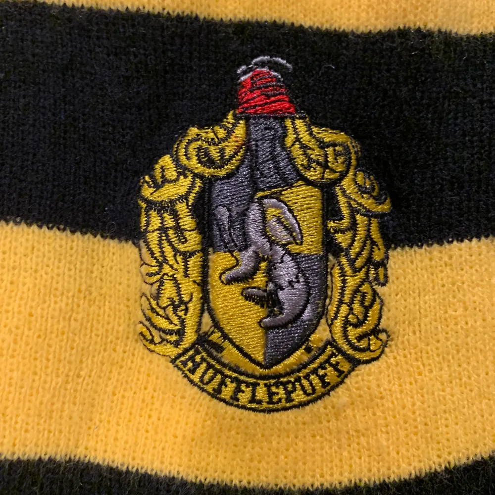 En randig halsduk i svart och gul med en embroiderad Hufflepuff logga. Oanvänd. Från Science Fiction Bokhandeln 80kr + frakt (cirka 50kr). Övrigt.