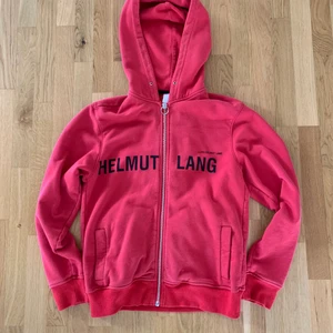 Helmut Lang hoodie  - Efterfrågad röd Helmut Lang hoodie. Storlek S men passar mer M. Bra skick förutom att jag bytt ut den man drar upp och ner dragkedjan med då den gick av. Snören finns sparade men har dragit ut dem.