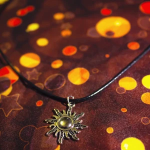 Halsband med solhänge - Handgjort (av mig) halsband med ett metallhänge i form av en snirklig sol. Halsbandet har en låsmekanism i metall högst upp. 🖤🌞