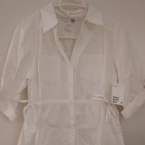 NY & oanvänd vit skjortklänning / blus. Storlek: 34/XS. Helt felfri. - NY och oanvänd vit skjortklänning/blus. Har 2 st bröstfickor, knäppning framtill, knyt i sidorna och reglerbar ärmlängd. Storlek: 34/XS.  Material: Bomull. Lappar/tags sitter kvar sedan inköpet. Helt felfri. FRAKT TILLKOMMER.