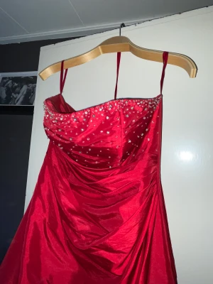 Elegant röd Balklänning  - Syuperfin röd balklänning i silky tyg, med stenar/kristaller över bysten.  Klänningen har inga axelband, det finns en sjal till, & i ryggen finns en dragkedja.  Kjolen är något vid nedtill, men känns relativt nätt att bära.  KOM MED rimliga BUD! 