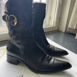 Svarta / mörkbruna stövletter / boots - Stövlar med dragkedja i svart (mörkt mörkt brunt) läder. Snygga detaljer med rem och spänne i guld/mässing. Gummisula. Storlek 41. Evita heter märket. Nypris 2000krz Skorna är gjorda i Italien. Sparsamt använda. Klacken är ca 3 cm. Ofodrade. 