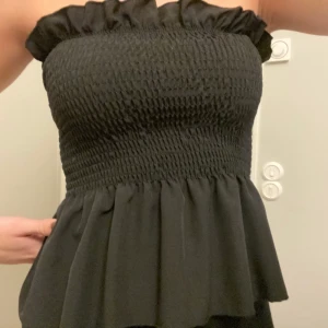 Svart topp - OANVÄND! Svart topp utan axelband. Smock med en liten volang nertill. Kommer tyvärr inte till användning. Köparen står för valfri frakt!