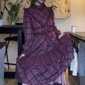 Vintage klänning - Lila/rödrutig vintage klänning från finska Merry Finn. Stängs med dragkedja i ryggen. Knytskärp i midjan. Volanger på hela kjolen, fodrad med tunt polyestertyg. 45% ull, 55% polyester. Perfekt höst/vinterklänning!  Lite nopprig pga materialet. US 8/36