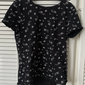 Svart Tukan Tröja - En svart t-shirt med vita tulpaner och blommor på. Den är gjord av bomull och mysigt, har inte används mycket.