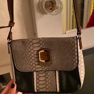 Axelremsväska  - En snygg axelremsväska som man kan justera i handen. Den är liten men praktisk, rymmer mobil, nycklar, plånbok och även mer prylar. Den är svart, vig och grå med lite bruna detaljer. Säljer för 250kr, köpte den för 599. 