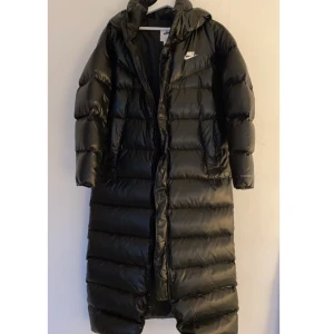 Nike vinterjacka  - En lång, varm och glansig vinterjacka från Nike köpt på Stadium i november 2021. Använd bara 2 gånger under den senaste vintern. Så gott som NY! Inga skador, inga slitningar. Säljer pga att den nu blivit för liten för att knäppa. 