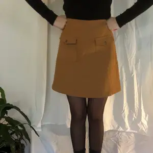Söt beige, fake-mocka-kjol, med fickor på vardera sida framtill. Kort modell.