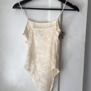 Body med spetsrygg • strl M - Beige body med spetsrygg i storlek M. Använd 1 gång så är i fint skick. Sitter som ett figurnära linne på överkroppen. Bilden visar baksidan av plagget. 