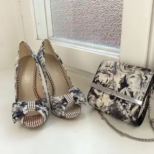 Snygga skor i blommigt mönster (svart/vit rosor) med rosett. Den ena skor har blivit lite ”färgad” på framdelen. Syns inte när skor är på. Passande liten väska följer med. 