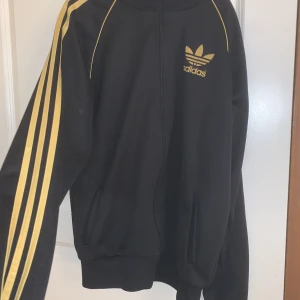 Adidas kofta - Den är i bra skick och den är svart med guld detalj och den är väldigt fin men stor. 