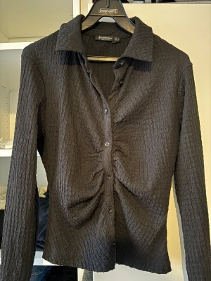 Svart skjorta från Stradivarius M - Svart tröja från Stradivarius i storlek M/S med krage och lång ärm. Materialet är krinklat och har en snygg rynkad effekt längs knappraden framtill. Skjortan har klassiska knappar och en figurnära passform som ger en fin form.