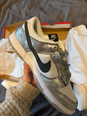 Nike Dunk Low Silver/Glitter sneakers - Nike Dunk Low sneakers i silver och glitter i storlek 41 köpta i USA. Kvar i kartong. Ovandelen är i mesh och glittrigt syntetmaterial, med grå snörning och vit mellansula. Yttersulan är ljusgrå och har klassiskt Dunk-mönster.                      Aldrig använda, säljer pågrund av att jag råkade köpa fel storlek 🩷 Nypris 2000kr 