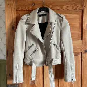 Beige mocka jacka med bälte - Snygg beige jacka i mocka från Zara med coola detaljer som bälte i midjan, dragkedjor på fickor och ärmar samt axelklaffar. Jackan har en croppad passform och klassisk krage, perfekt för dig som gillar en edgy stil. Ny utan prislapp. Används bara en gång. 