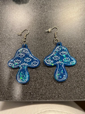 Blå svampformad örhängen med glitter - Unik blå örhängen i svampform med glittriga detaljer och vita ögonmönster. Örhängen har en cool, konstnärlig vibe. Perfekt för dig som gillar statement-plagg och vill sticka ut med din stil.