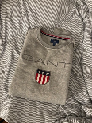 Grå sweatshirt från GANT med emblem - Säljer en grå sweatshirt från GANT i storlek S. Tröjan har ett broderat GANT-emblem med stjärnor och ränder i rött, vitt och blått på bröstet samt GANT-logga ovanför. Rund halsringning och mjukt bomullsmaterial. Perfekt för chill dagar.