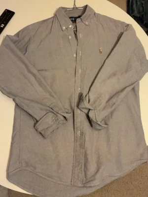 Grå skjorta från Ralph Lauren - Säljer en grå skjorta från Ralph Lauren, modell Custom Fit. Skjortan har klassisk krage, långa ärmar och knappar framtill. Materialet är mjuk bomull och det finns en liten broderad logga på bröstet. Perfekt för dig som gillar stilrena plagg med snygg passform. Modell: 175cm