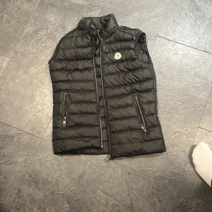 Svart dunväst från Moncler - Säljer en svart dunväst från Moncler med horisontella sömmar och två dragkedjefickor framtill. Västen har hög krage och klassisk Moncler-logga på bröstet. Perfekt att slänga över en hoodie eller tröja när det är kyligt ute.