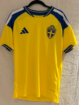 2026 Sverige tröja - Säljer en svensk landslagströja i stark gul färg med blå och vita detaljer. Tröjan har broderat landslagsemblem och Adidas-logga på bröstet, samt texten 'Sverige' på ryggen. Tillverkad i lätt polyester med Climalite-teknologi.