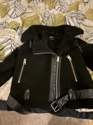 Svart teddyjacka med bälte från Zara - Säljer en svart teddyjacka från Zara med feta dragkedjor, bälte i skinnimitation och mjukt teddyfoder. Jackan har stora fickor med dragkedja och detaljer i skinnimitation på ärmar och nederkant. Perfekt för dig som vill ha en cool och varm look. Fraktar enbart via post nord!!!!