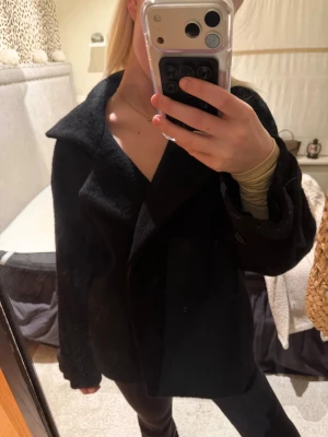 Svart ullkappa med bred krage Arket  - Säljer en svart kappa i ull från Arket med bred krage och dubbelknäppning framtill. Jackan har en loose passform och långa ärmar, perfekt för höst och vinter. Stilren och enkel design som funkar till många outfits.