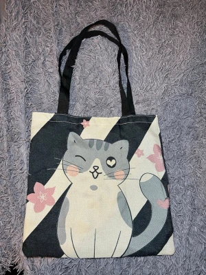 Shoppingväska med kattmotiv - Superfin shoppingväska med ett gulligt kattmotiv som blinkar och har ett hjärta i ögat. Väskan har svarta handtag och är dekorerad med rosa blommor mot en beige och svart bakgrund. Perfekt storlek för böcker eller laptop, och riktigt söt design.