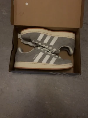 Grå Adidas Campus sneakers - Säljer ett par grå Adidas Campus sneakers med klassiska vita ränder på sidan och vit sula. Skorna har snörning och är gjorda i mjuk mocka med en platt sula. Perfekta för dig som gillar stilrena och tidlösa sneakers.