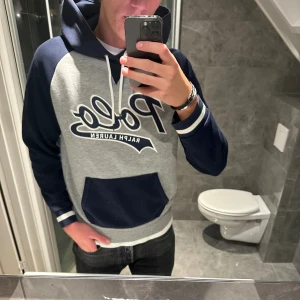 Grå och marinblå hoodie Polo Ralph Lauren - Säljer en snygg hoodie från Polo Ralph Lauren i grått och marinblått med stor logga framtill. Tröjan har huva med snörning, känguruficka och ribbade muddar. Hoodien är k riktigt bra skick. Jag är 188cm på bilden. Hör av er vid funderingar;)