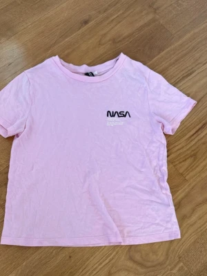 Ljusrosa NASA t-shirt från DIVIDED - Säljer en ljusrosa t-shirt från DIVIDED i storlek XS med svart NASA-tryck på bröstet. T-shirten har rund hals och korta ärmar, tillverkad i mjuk bomull. Perfekt för dig som gillar rymdtema och vill ha en chill look.