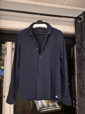 Mörkblå skjorta från Rose & Born - Snygg mörkblå skjorta från Rose & Born med klassisk krage och knappar framtill. Skjortan har lång ärm och är skräddarsydd för en stilren passform. Perfekt för dig som gillar enkel och clean stil.