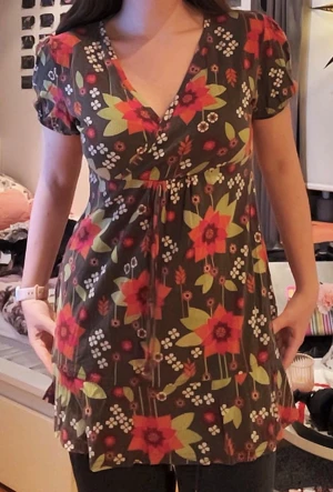 Blommig kortklänning med v-ringning - Supersöt kortklänning med v-ringning och blommigt mönster i rosa, rött, grönt och vitt på mörk botten. Klänningen har korta puffärmar och volang nertill. Perfekt för dig som gillar färg och mönster! Materialet känns mjukt och skönt mot huden.