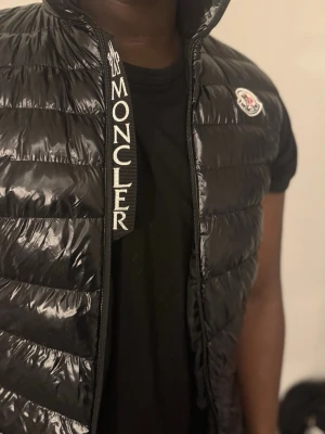 Svart dunväst från Moncler - Säljer en svart, glansig dunväst från Moncler med broderad logga på bröstet och tydlig Moncler-text på insidan av dragkedjan. Västen har quiltad design och är tillverkad i polyester med dunfyllning. Perfekt för lager-på-lager under kyliga dagar. Är lite sönder på innerfickan men kan troligtvis fixas om man syr. I storlek s men passar M/L