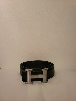 Svart bälte med ett stort H i mitten. - Bilden visar ett svart bälte med ett stort H-format spänne i silverfärg. Får med box som jag köpte med bältet.