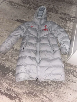 Ljusgrå lång dunjacka Liverpool Nike - Lång, ljusgrå dunjacka från Nike i samarbete med Liverpool FC. Jackan har huva, två dragkedjeförsedda fickor och röda loggor på bröstet. Perfekt för kalla dagar med sin pufferstil och quiltade sektioner. Snygg och sportig look.