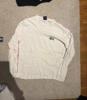 Vit långärmad topp från Stüssy - Vit långärmad topp från Stüssy med svart logotyptryck både fram och stort bak. Klassisk streetwear-look med rund hals och mjukt bomullsmaterial. Perfekt för dig som gillar enkel men snygg design och vill ha något som sticker ut med loggan.