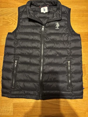 Svart dunväst från US Polo Assn - Svart dunväst från US Polo Assn med horisontella quiltade paneler och två fickor med dragkedja. Västen har ståkrage och USPA-logga broderad på bröstet. Perfekt för lager-på-lager och snygg till grisch stilen.😃
