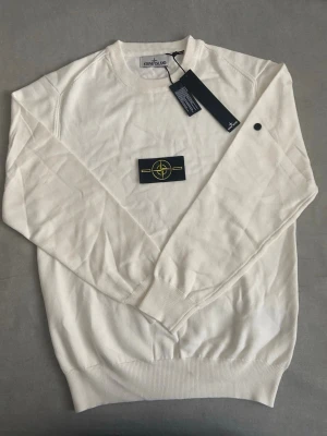 Vit Stone Island tröja M - Vit långärmad tröja från Stone Island i mjuk bomull. Klassisk rund hals och ribbade muddar. Ikonisk Stone Island patch på bröstet i svart och gult. Perfekt för dig som gillar stilrena och exklusiva plagg.