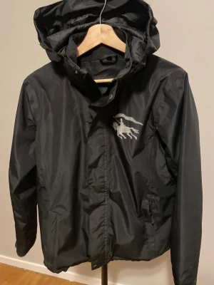 Svart Burberry Vindjacka - Svart vindjacka med huva och coolt tryck av en ryttare med flagga på bröstet. Jackan har dragkedja framtill och praktiska fickor. Perfekt för dig som gillar stilrena och sportiga plagg med en unik detalj.