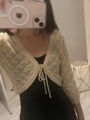 Beige stickad kofta med knyt - Säljer en beige/guldig stickad kofta/bolero med snyggt mönster och knyt framtill. Koftan är croppad och har trekvartsärm, perfekt att slänga över en topp. Det stickade materialet har ett luftigt, dekorativt mönster som ger en chill vibe. jag är 160cm