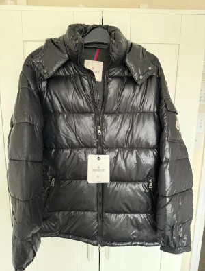 Svart pufferjacka från Moncler - Svart pufferjacka från Moncler med glansig finish och klassisk logga på ärmen. Jackan har hög krage, dragkedja framtill och praktiska fickor med dragkedja. Perfekt för kalla dagar och har en riktigt snygg streetstil.