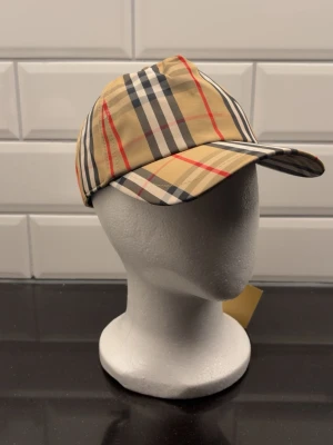 Rutig Burberry keps i beige och svart - Snygg keps från Burberry med klassiskt rutigt mönster i beige, svart, vitt och röda detaljer. Justerbar baktill med svart spänne och Burberry London England-märke. Perfekt accessoar för dig som gillar ikonisk design och streetstyle.