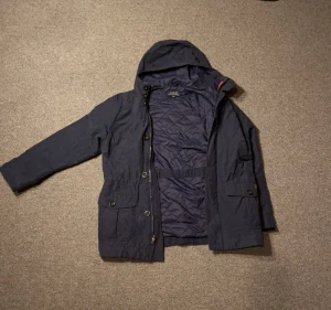 Mörkblå parkajacka från Polo Ralph Lauren - Parkajacka i mörkblått från Polo Ralph Lauren med huva, quiltat foder och stora fickor med knappstängning. Jackan har dragkedja och knappar framtill, samt justerbar midja för snygg passform. Perfekt för kyliga dagar och har en clean, urban vibe. Storlek M 10-12