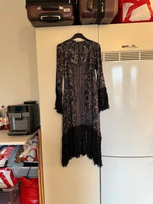 Svart mönstrad långklänning Odd Molly - Unik långklänning från Odd Molly i svart och grått med detaljerat mönster och fransar vid ärmslut och nederkant. Klänningen har v-ringad hals och är tillverkad i 100% viskos, vilket gör den mjuk och följsam. Perfekt för dig som gillar boho-vibe och snygga detaljer. Storlek 1 som enligt deras storleksguide motsvarar en S. men är rymlig i storlek.  Mycket fint skick, jag hittar inga synliga anmärkningar eller defekter. 