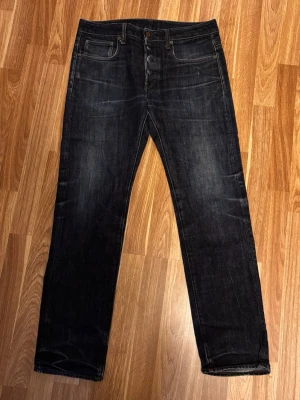 Mörkblå jeans från G-Star RAW - Snygga mörkblå jeans från G-Star RAW med klassisk femficksdesign och bruna detaljer på bakfickan. Jeansen har raka ben och orange kontrastsömmar som ger en schysst look. Perfekt för dig som gillar stilrena och tidlösa jeans.