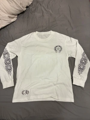 Vit långärmad Chrome Hearts tröja - Vit långärmad tröja från Chrome Hearts med stora svarta tryck på ryggen och bröstet, samt detaljerade grafiska mönster längs båda ärmarna. Tröjan har rund hals och är gjord i mjuk bomull. Perfekt för dig som gillar streetwear och unika designer.