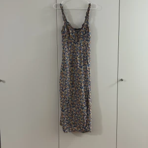 Blommig långklänning från F&B - Säljer en långklänning från F&F i storlek M med blommigt mönster i blått, orange och vitt. Klänningen har smock upptill, breda axelband och en slits framtill. Materialet känns som bomull och är lätt och luftigt, perfekt för varma dagar.
