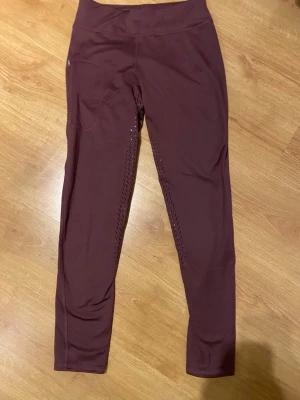 Vinröda leggings från Domino, strl 40 - Snygga vinröda leggings från Domino i storlek 40. Tillverkade i stretchigt material med 86% polyester och 14% elastan. De har hög midja och ventilerande håldetaljer längs benen, perfekta för träning eller chill. Supersköna och sitter tight.